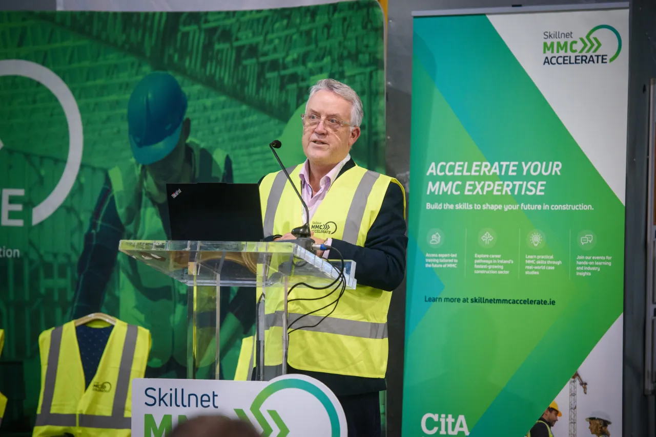 Person in safety vest speaking at podium with green event banner in background.