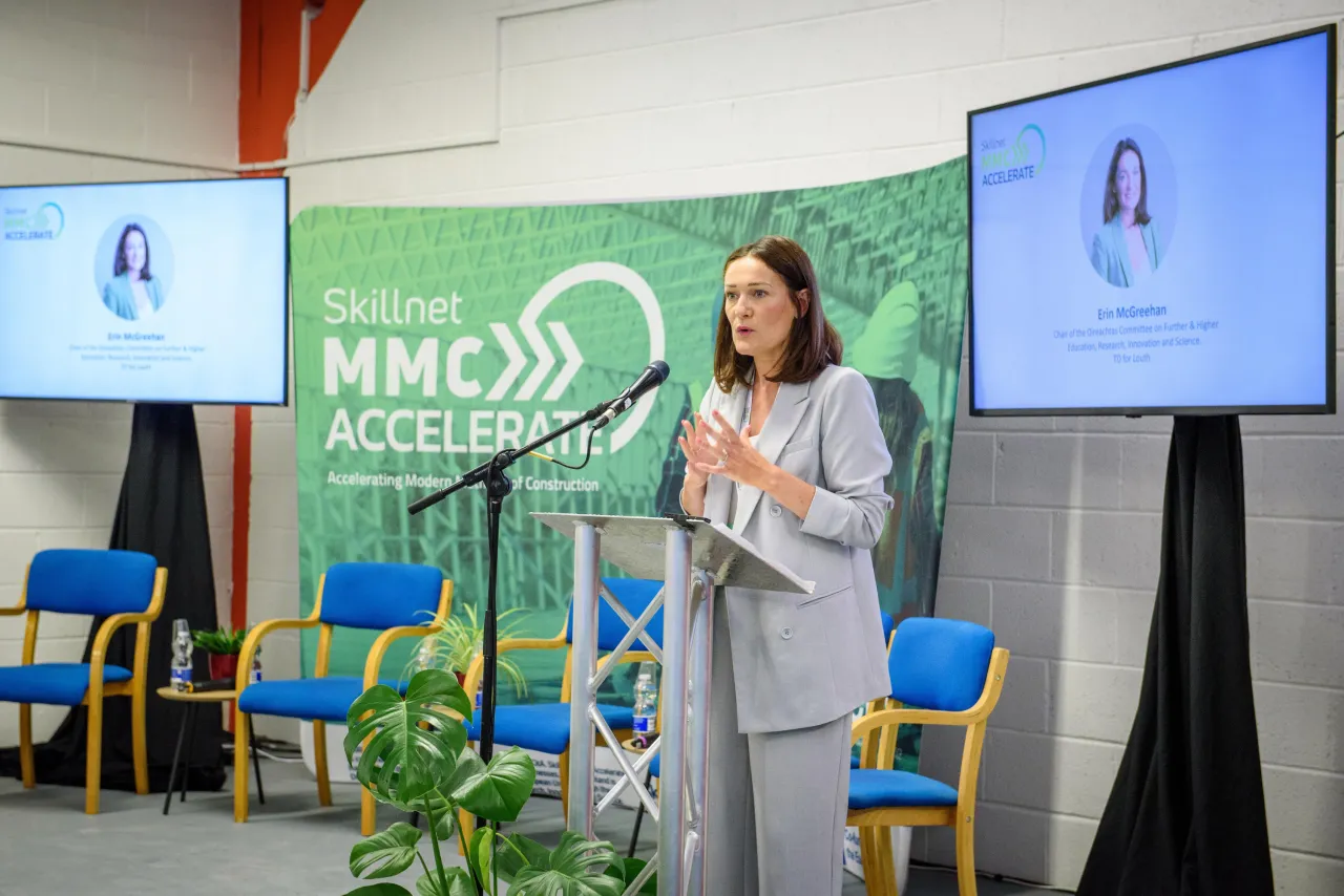 Woman presenting on stage with "NYC+" backdrop, blue chairs, and video screens.