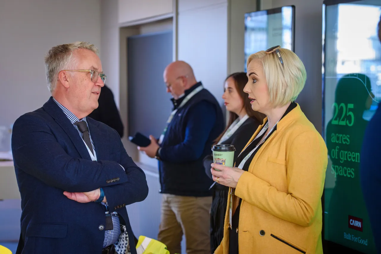 Three people in conversation at an indoor event, with a person wearing a yellow blazer gesturing while speaking.