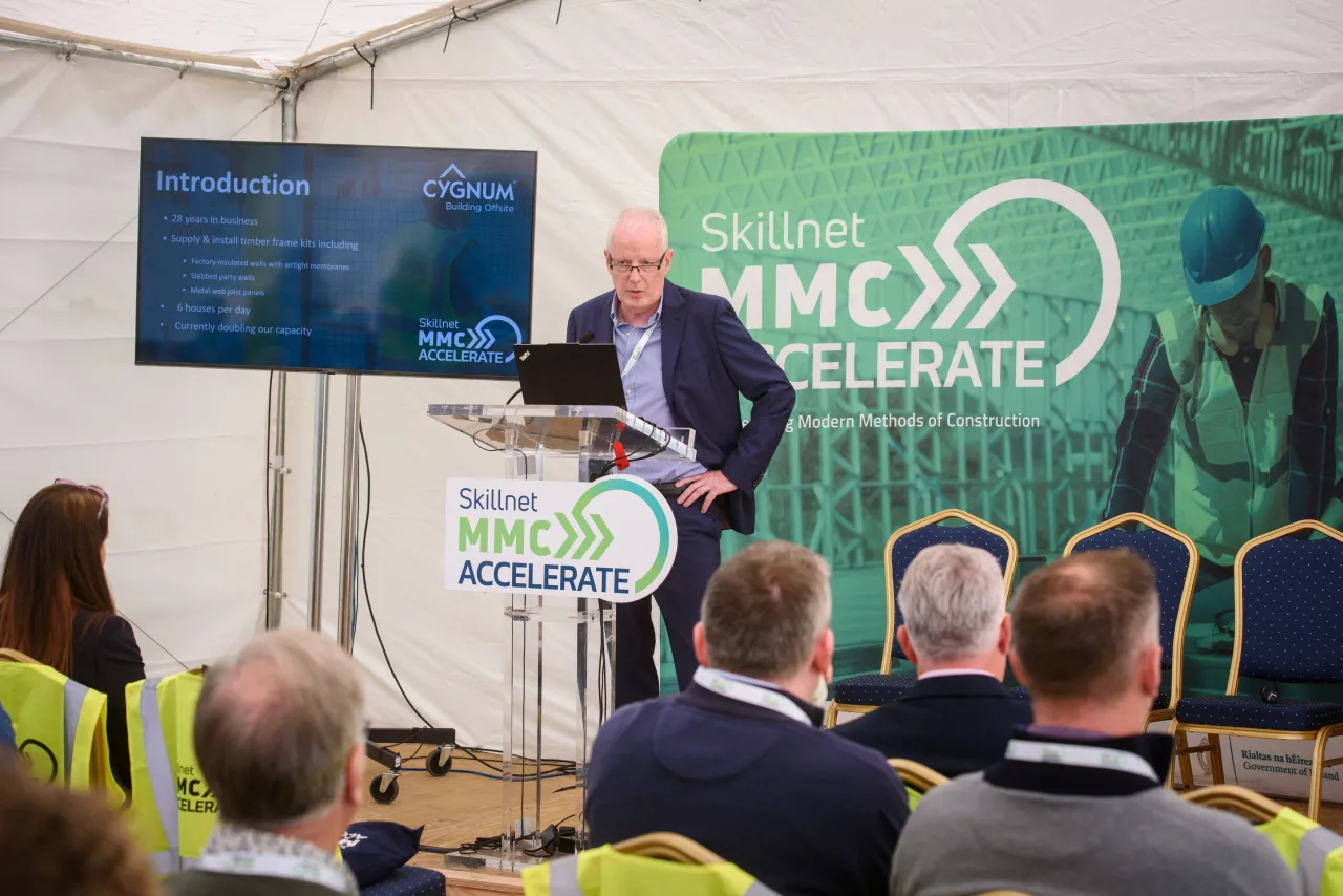 A speaker at a podium presenting to an audience in a white tent with event banners visible.