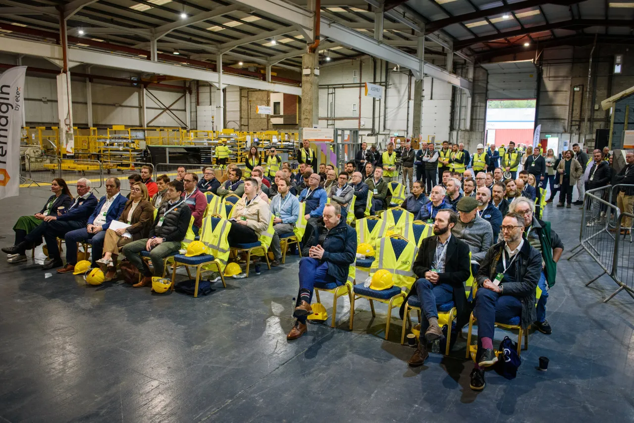 A large group of workers in safety vests gathered inside an industrial manufacturing facility.
