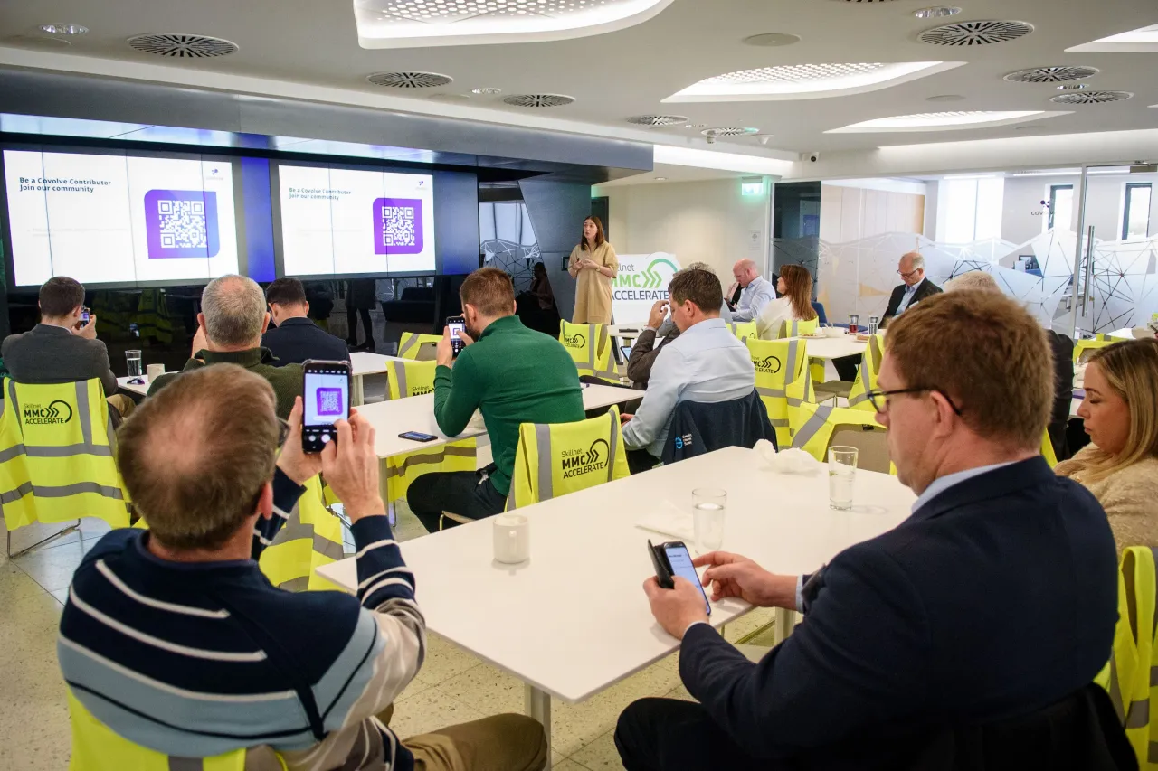 People working at tables in a modern office space with green chairs and digital displays on the walls.
