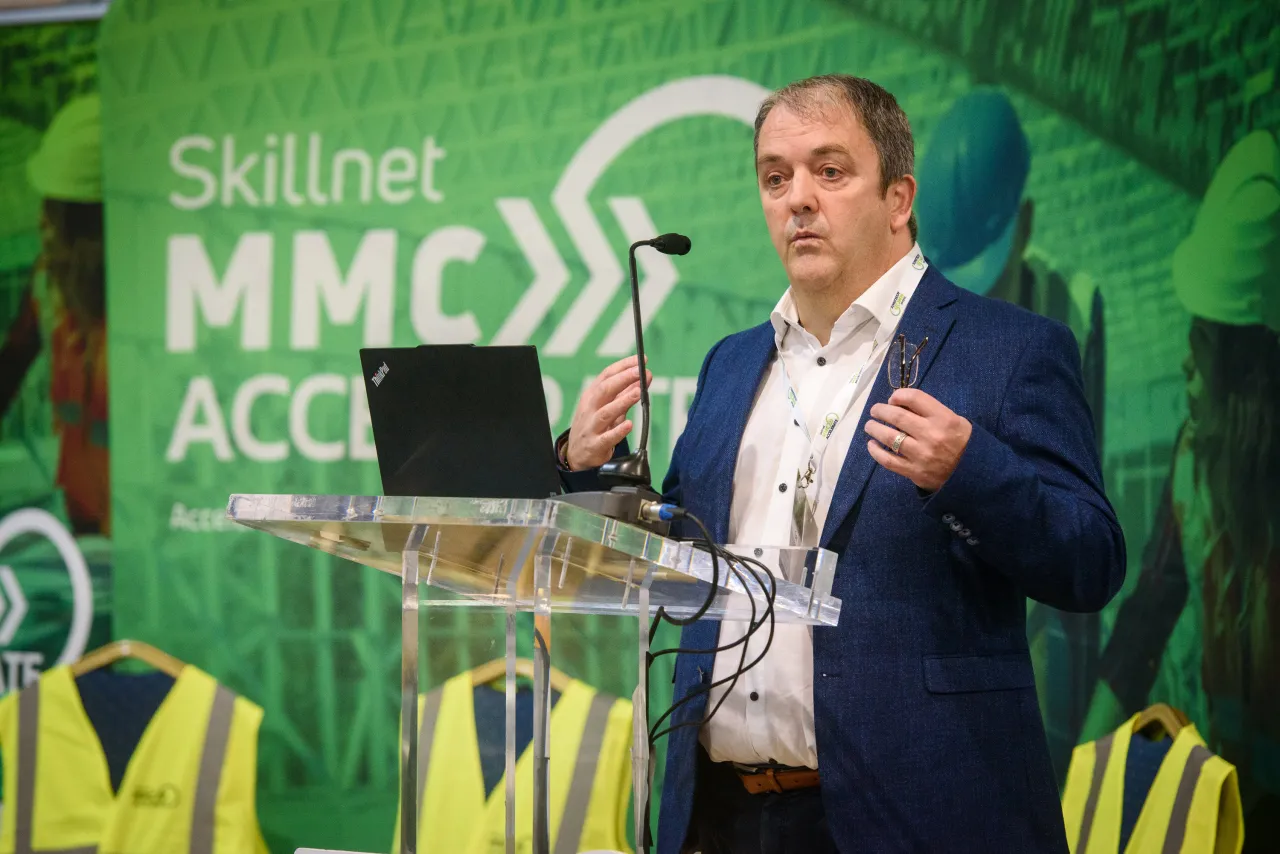 A speaker in a business suit at a presentation with green "Skillnet MMC ACCC" backdrop