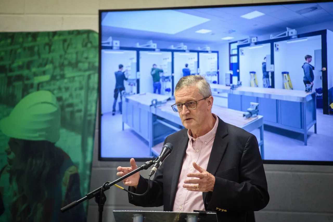 A person seated at a desk with microphone, with a large screen showing a video conference in the background.