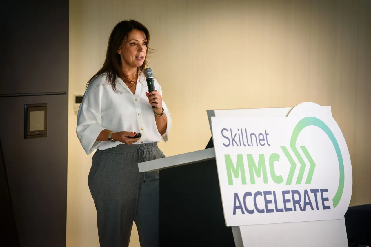 Woman in business attire speaking next to a sign with "Solve, Innovate, Accelerate" text.