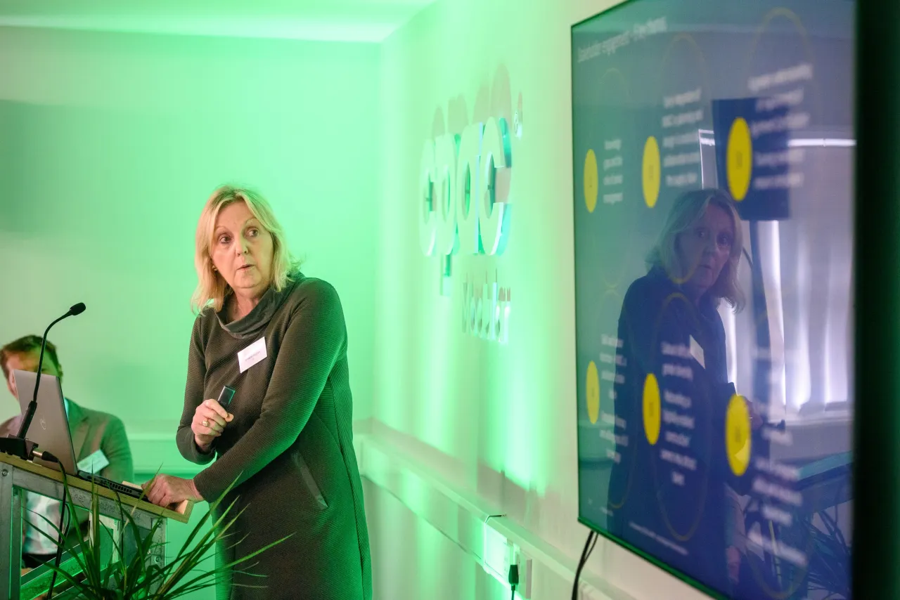 Person in green top performing music with electronic equipment and presentation screens in background.