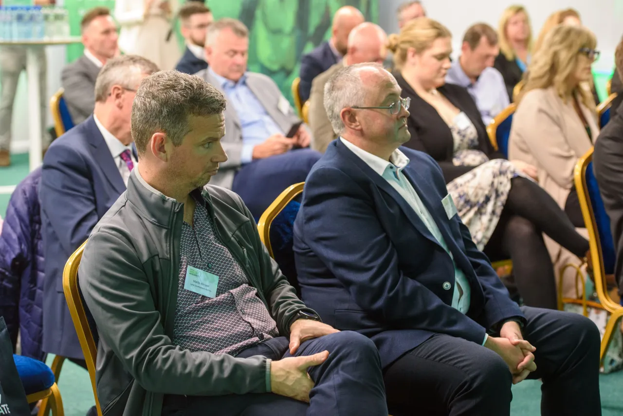 People seated at a business conference or presentation, focused on a speaker off-frame.