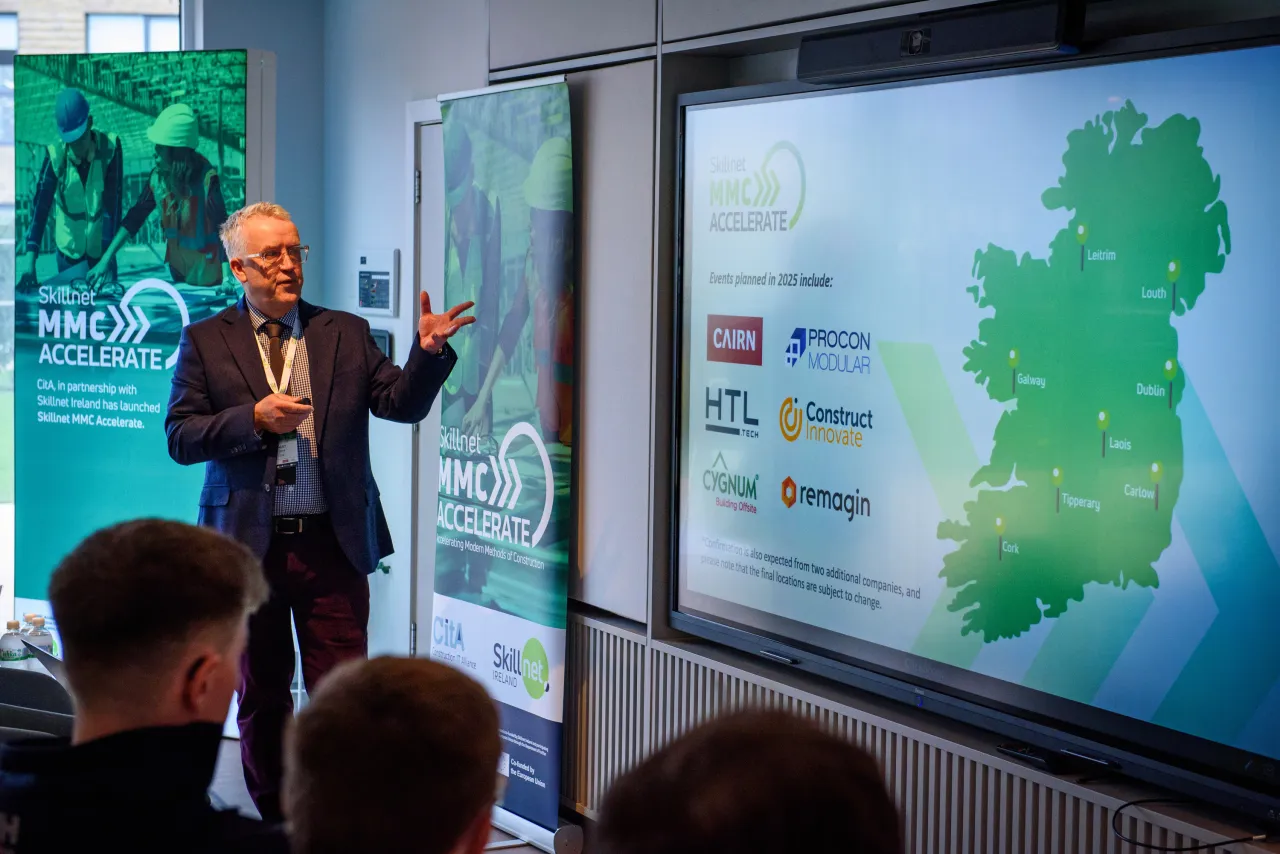 Person in formal attire presenting next to a screen showing a green map of Ireland.
