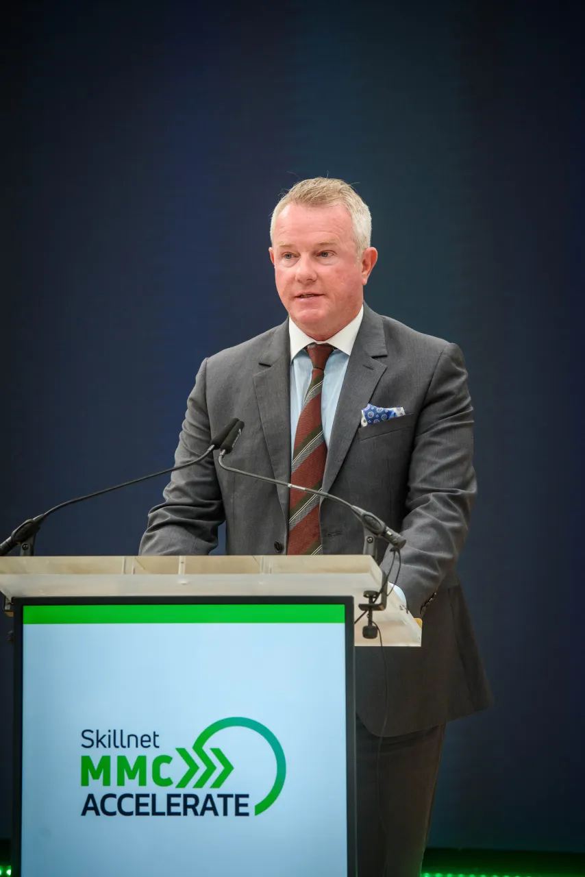 A person in formal suit speaking at a podium with the MHC Accelerate logo displayed.