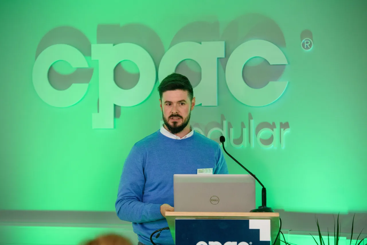 Person in blue sweater standing at a podium with laptop against green CPAC backdrop.
