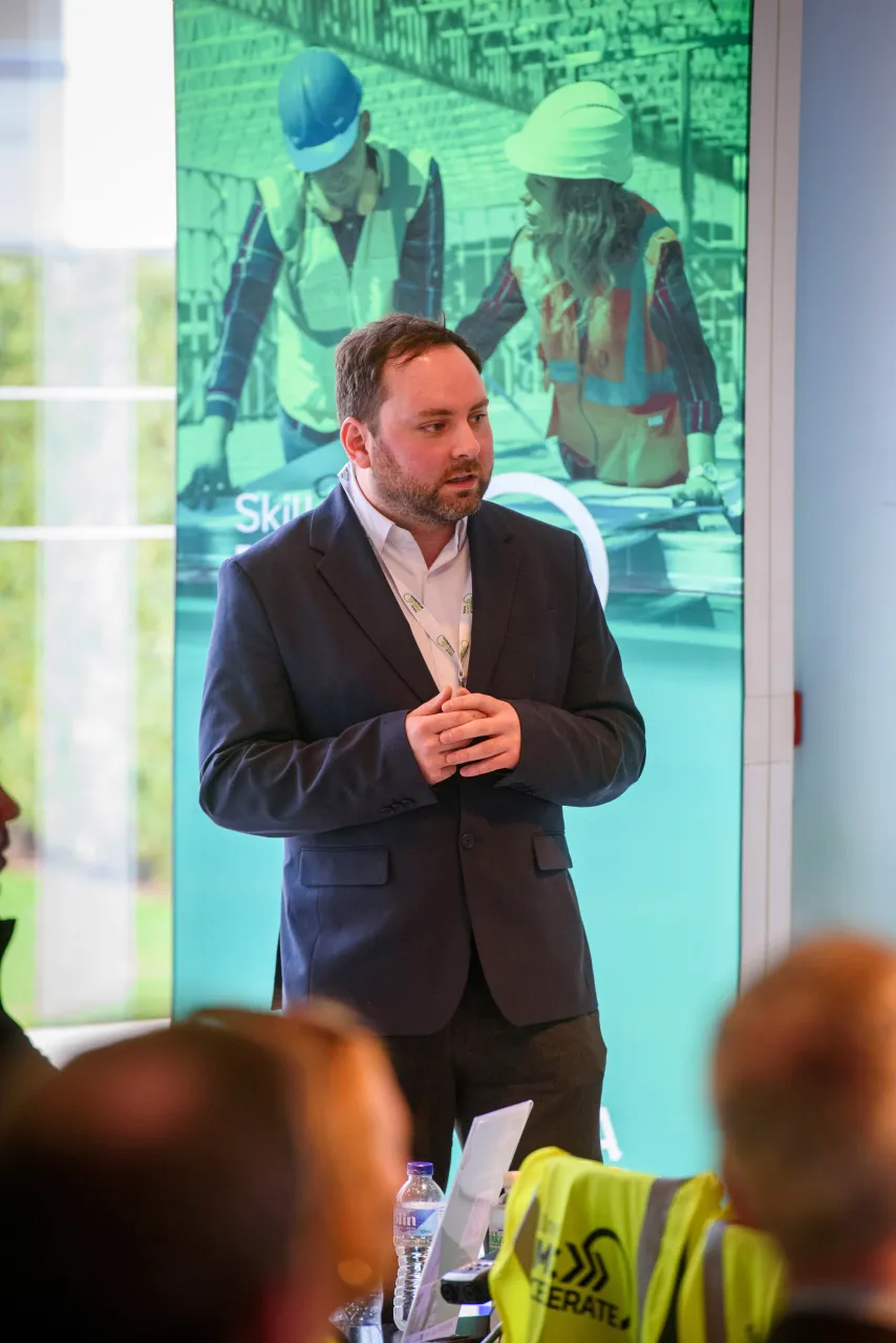 A man in a dark suit speaking at an event with a green display screen in the background.