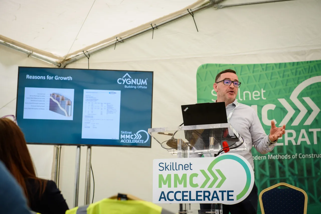 A person presenting at a small workshop or event with digital screens and green banners visible.