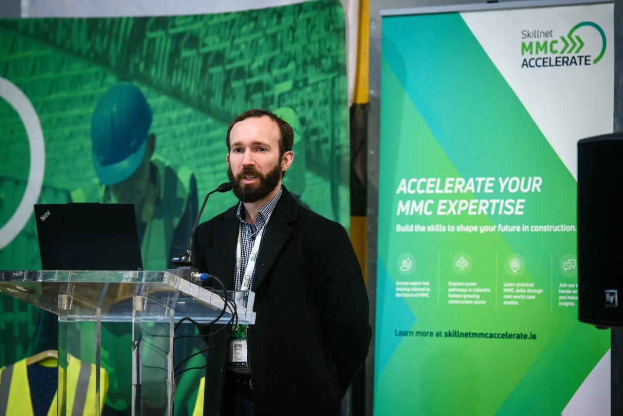 A person in business attire speaking at a podium with green backdrop and event signage.