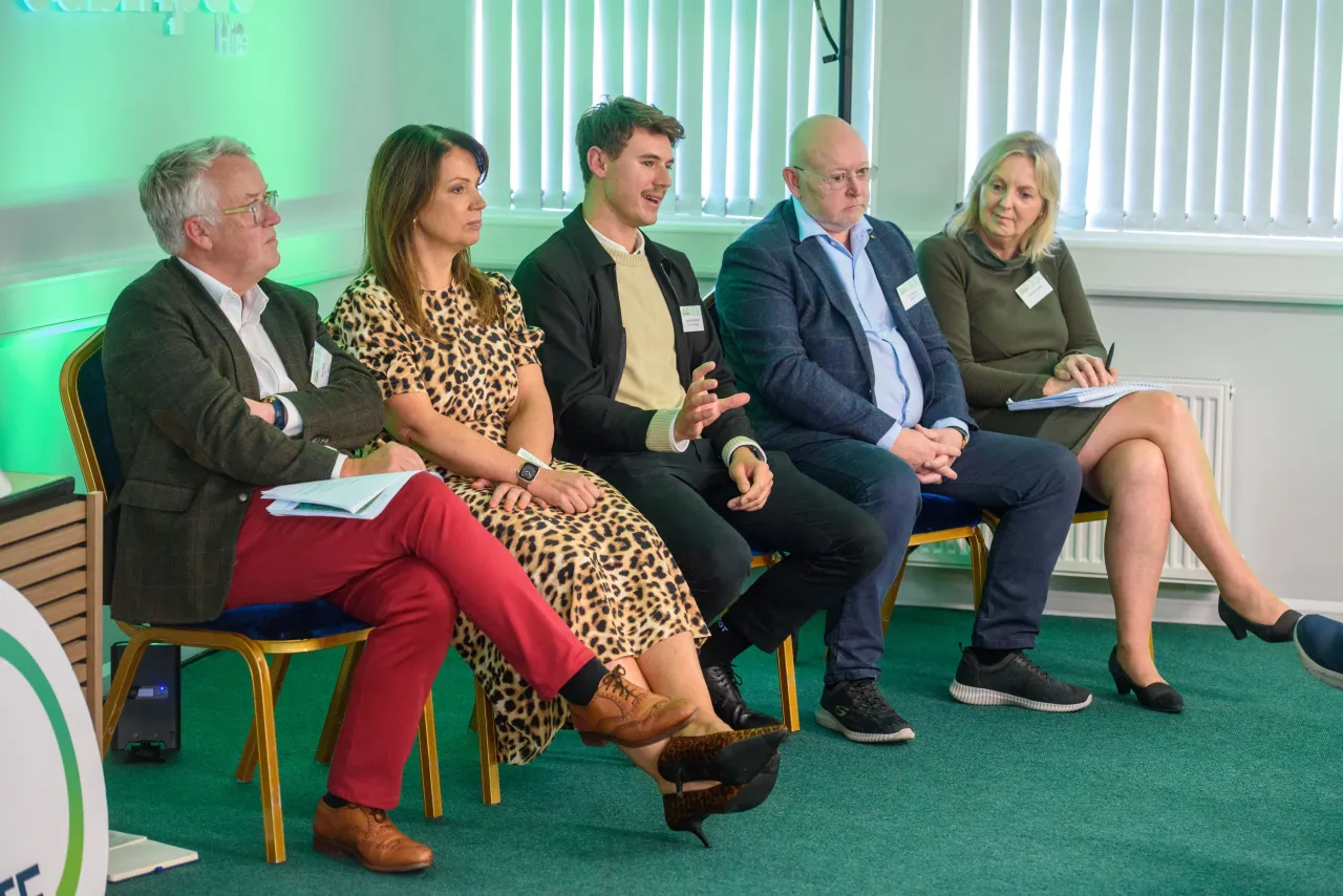 A panel of five people seated in chairs during a discussion event with green backdrop lighting.