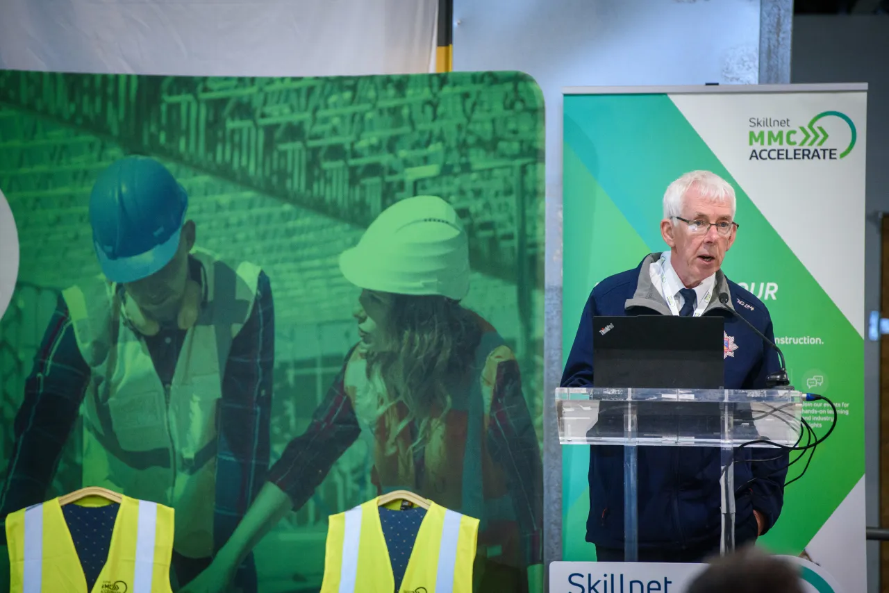 Speaker at a podium with construction workers visible on a green backdrop display.