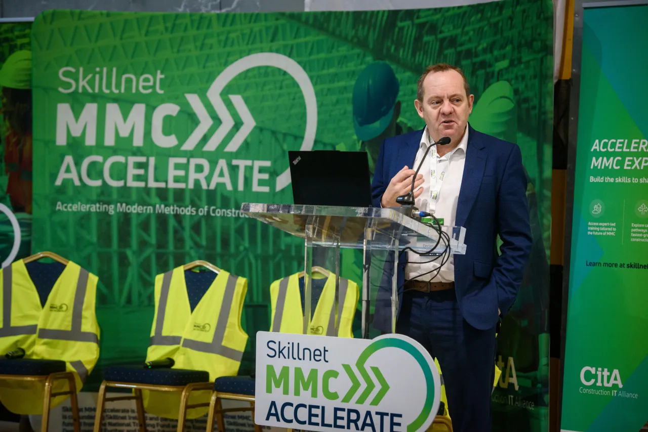 Person in formal attire presenting at MMC Accelerate event with green backdrop and safety vests.