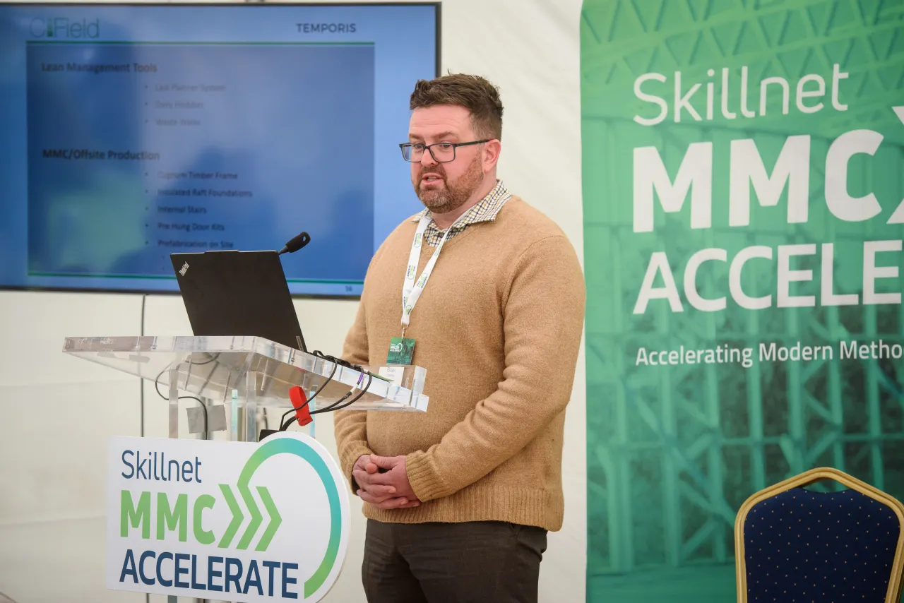 A person in a tan sweater presenting at a podium with Skillnet MMC Accelerate banners behind.