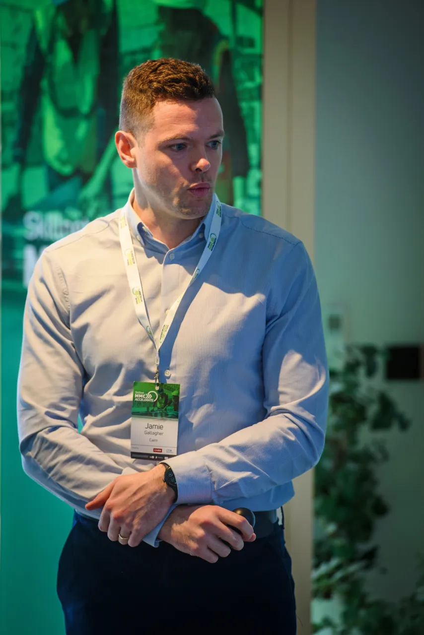 Young man in light blue shirt with name badge standing against green backdrop.