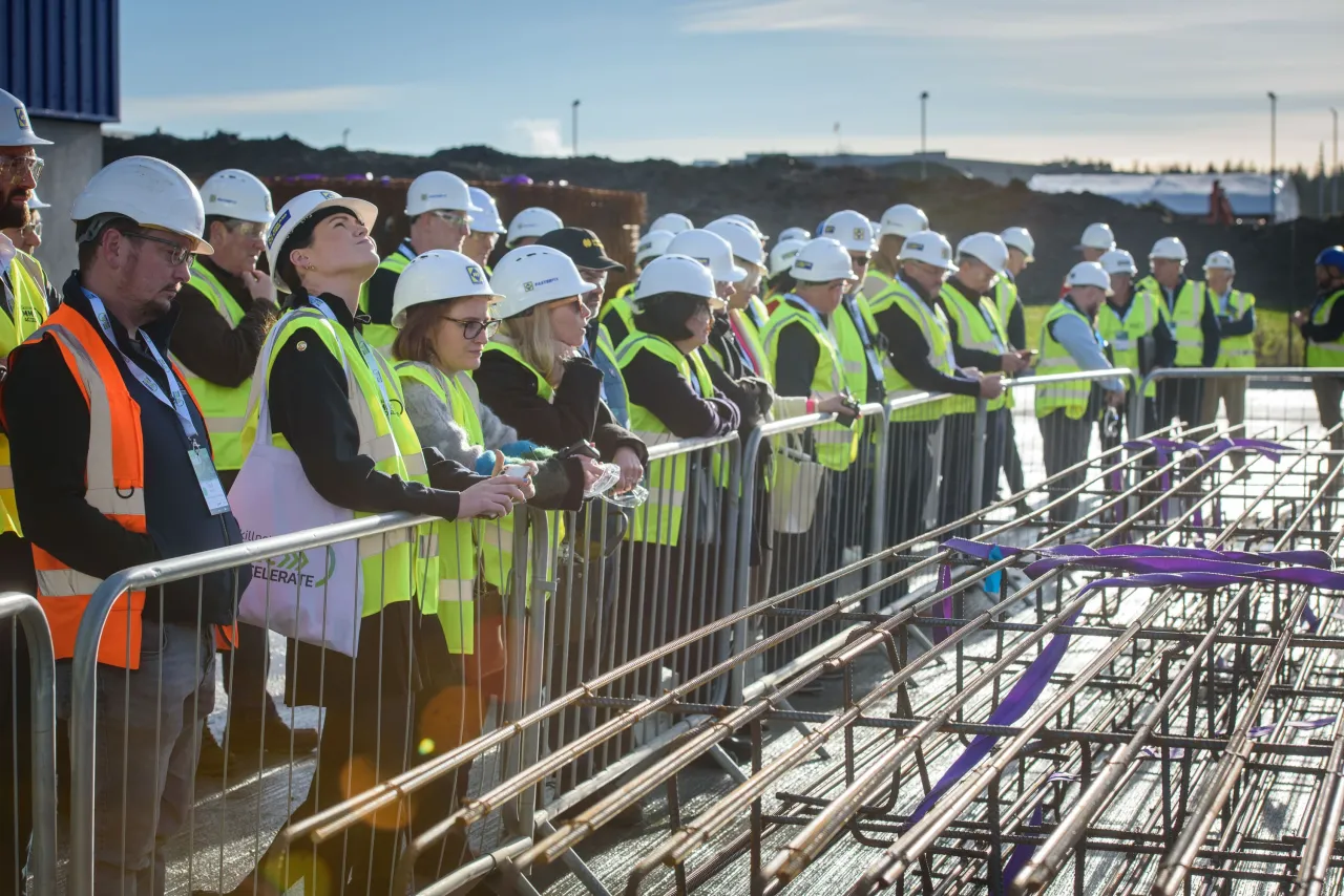 Group of construction workers in hard hats and safety vests standing along a railing at a construction site.