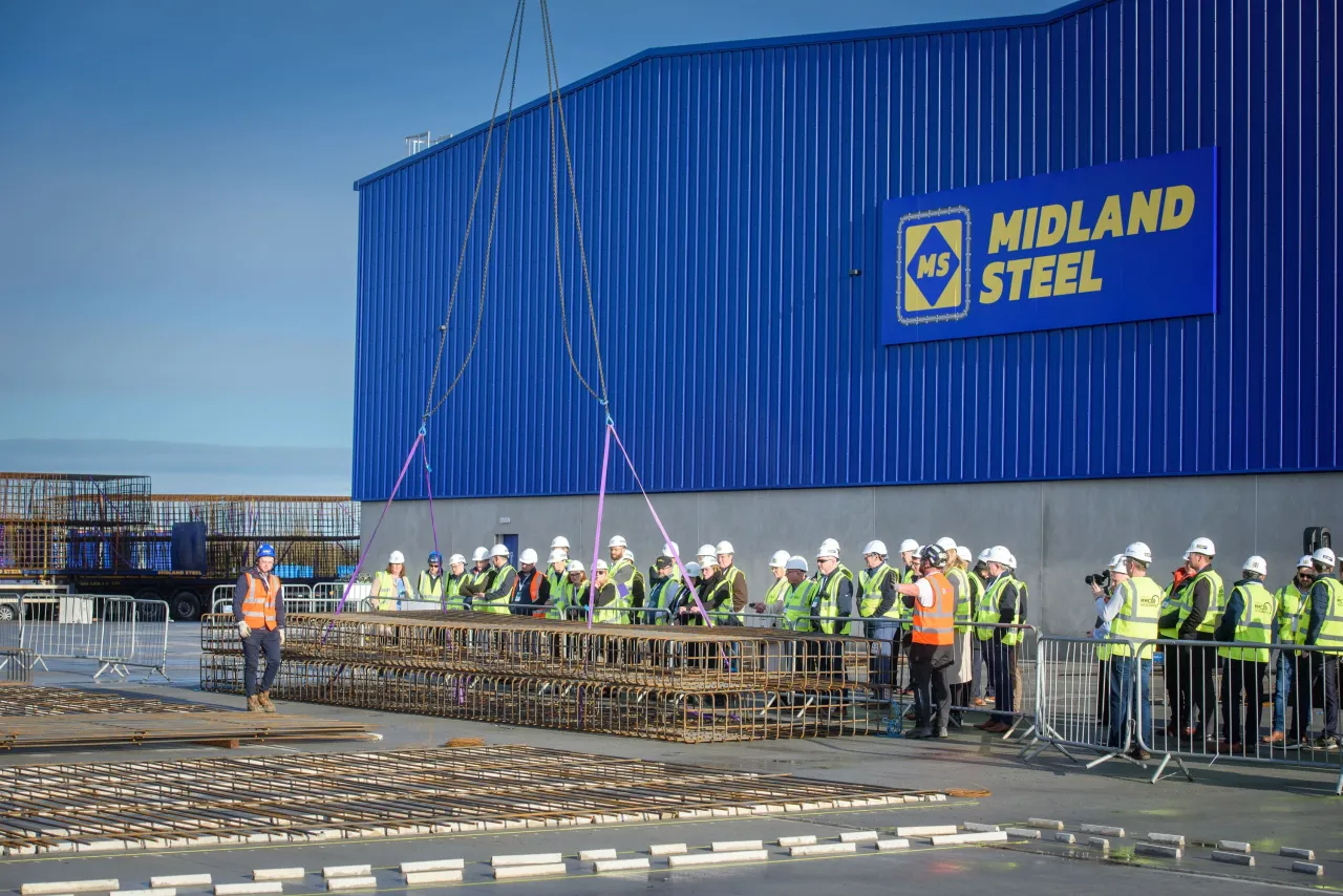 A group of workers in safety vests gathered outside a large blue Midland Steel facility building.