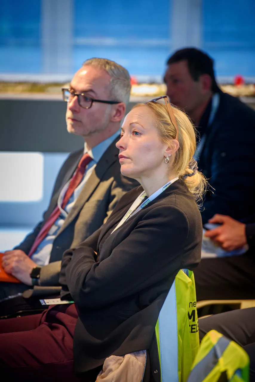 Business professionals sitting in a conference, woman in foreground wearing a dark suit jacket.