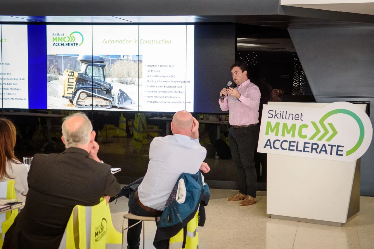 A presenter speaking at a business meeting with people seated at tables and a "Business Excellence" sign visible.