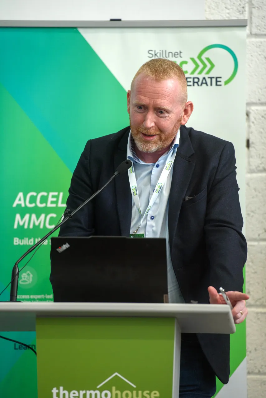Person in a suit speaking at a podium with a green backdrop featuring a logo.