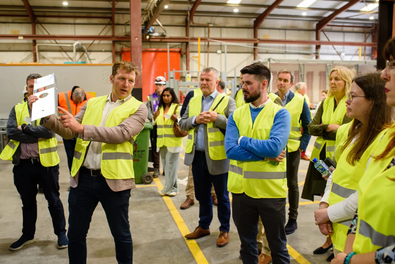 Group of people in yellow safety vests gathered in a warehouse for a tour or training session.