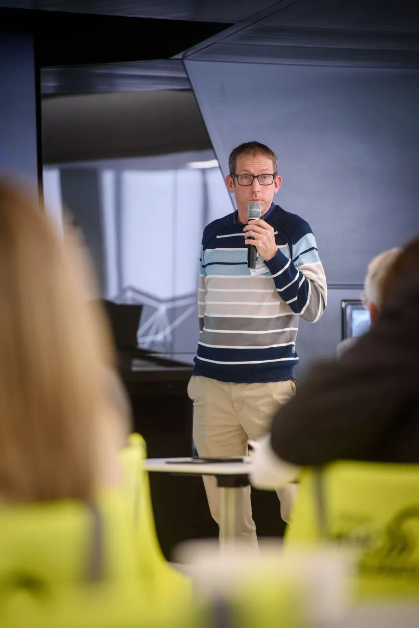 Person in striped shirt speaking at a podium to an audience in a classroom setting.