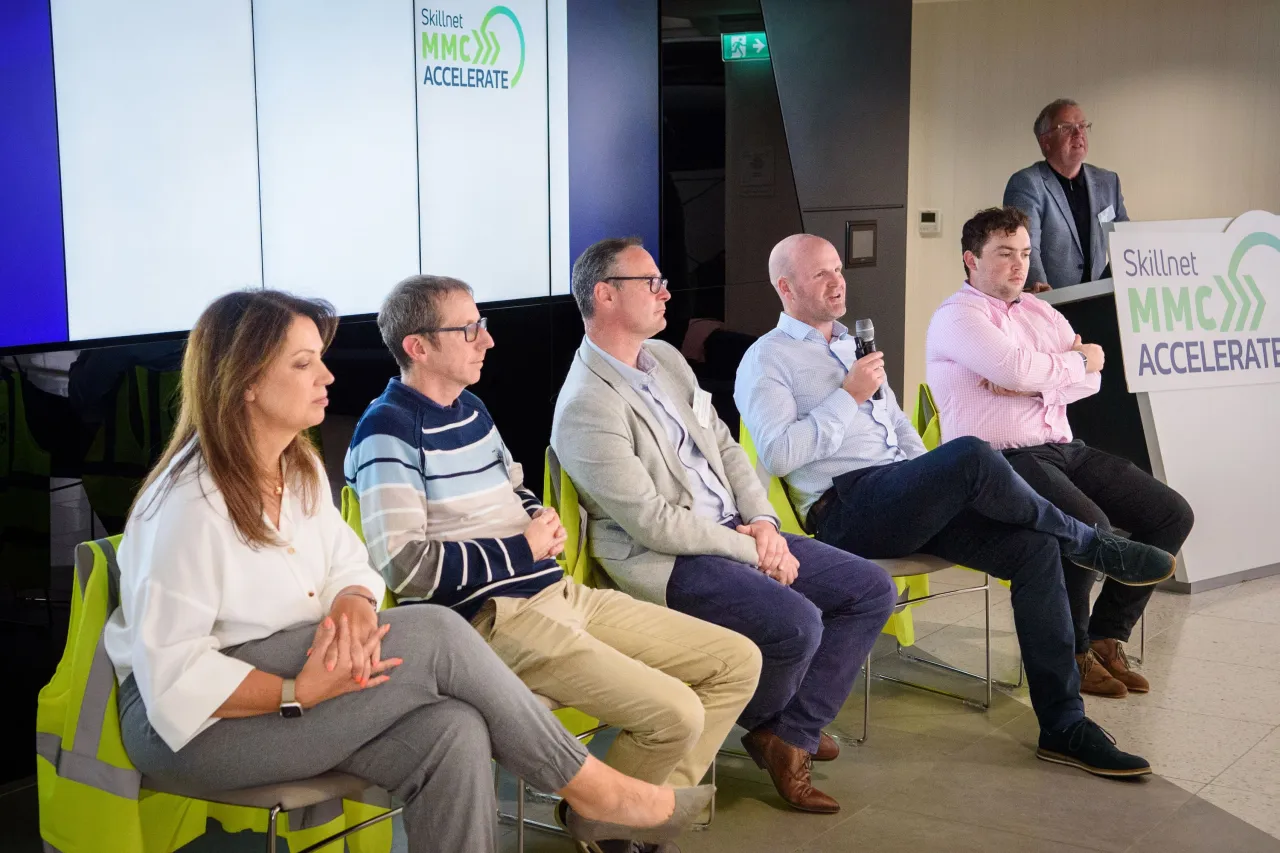 Panel discussion with people seated in green chairs on a stage with presentation screen visible.
