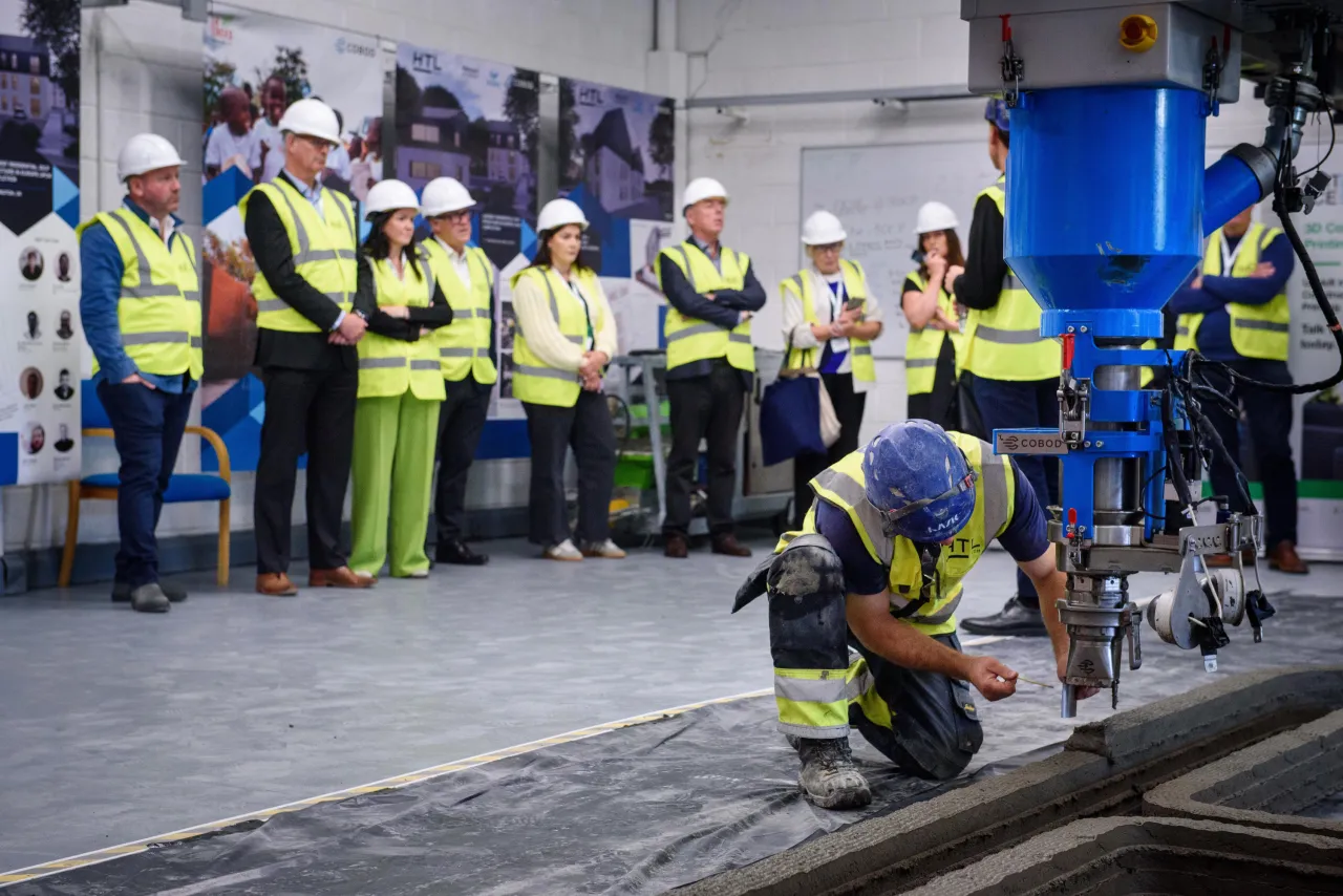 Construction workers in safety vests and helmets gather around equipment on a factory floor.