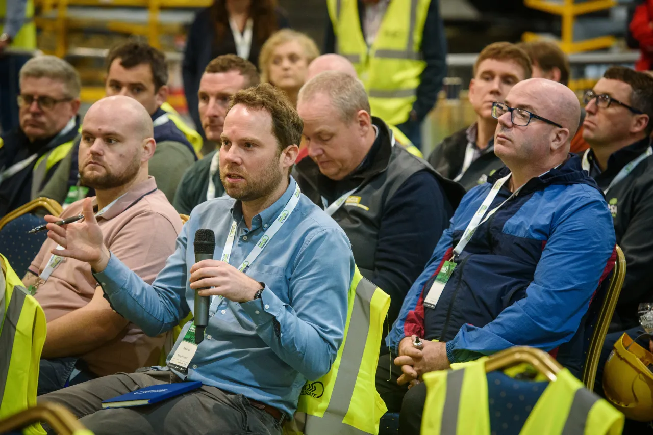 Men in safety vests seated at a business or training event, some wearing high-visibility clothing.