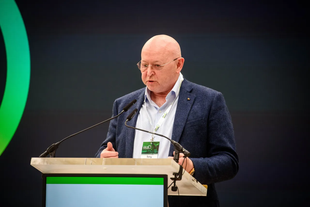 Person in a dark suit speaking at a podium with a green design element visible.