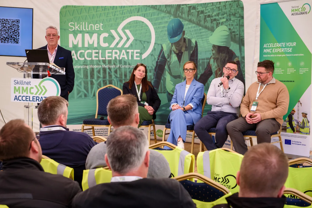 A panel discussion at a business event with speakers seated on stage and audience members visible.
