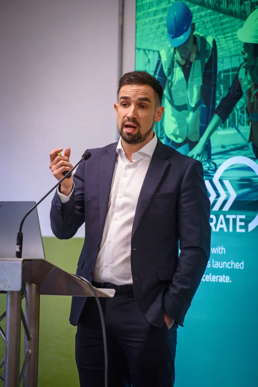 Man in suit speaking at podium with green conference banner visible behind him.