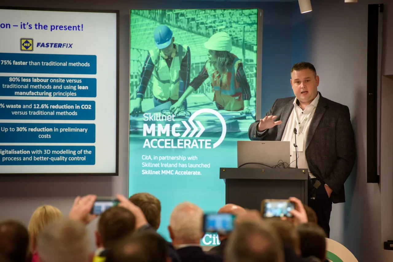 A presenter speaking at a business event with "INNOVATE NOW" displayed on a large screen behind him.