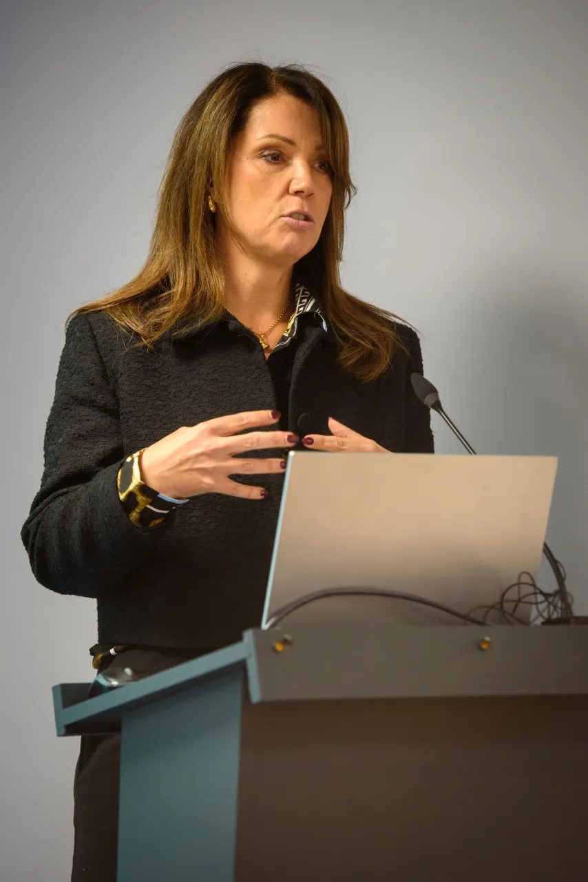 A person in a dark blazer speaking at a podium with a laptop.