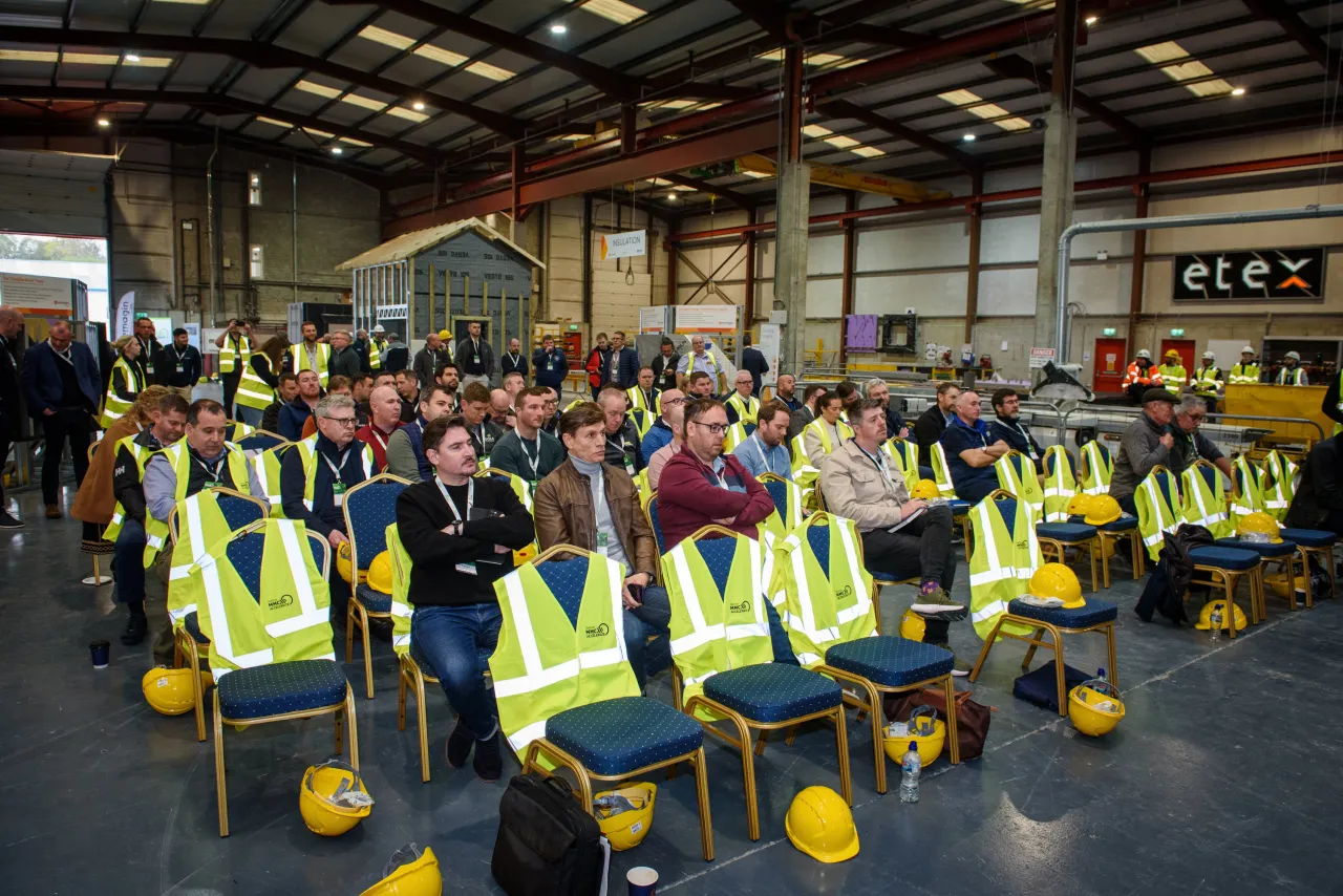 Workers in yellow safety vests and hard hats seated at a manufacturing facility meeting.