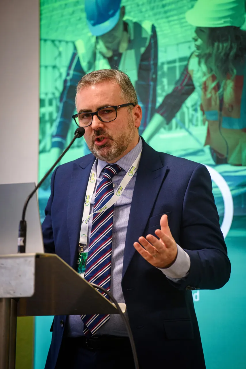 A man in a blue suit and tie speaking at a podium with a green presentation screen behind him.