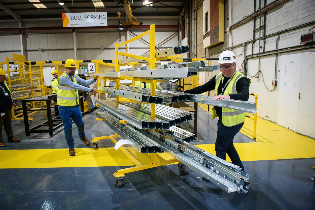 Workers in yellow safety vests and hard hats handling large aluminum beams in a warehouse facility.
