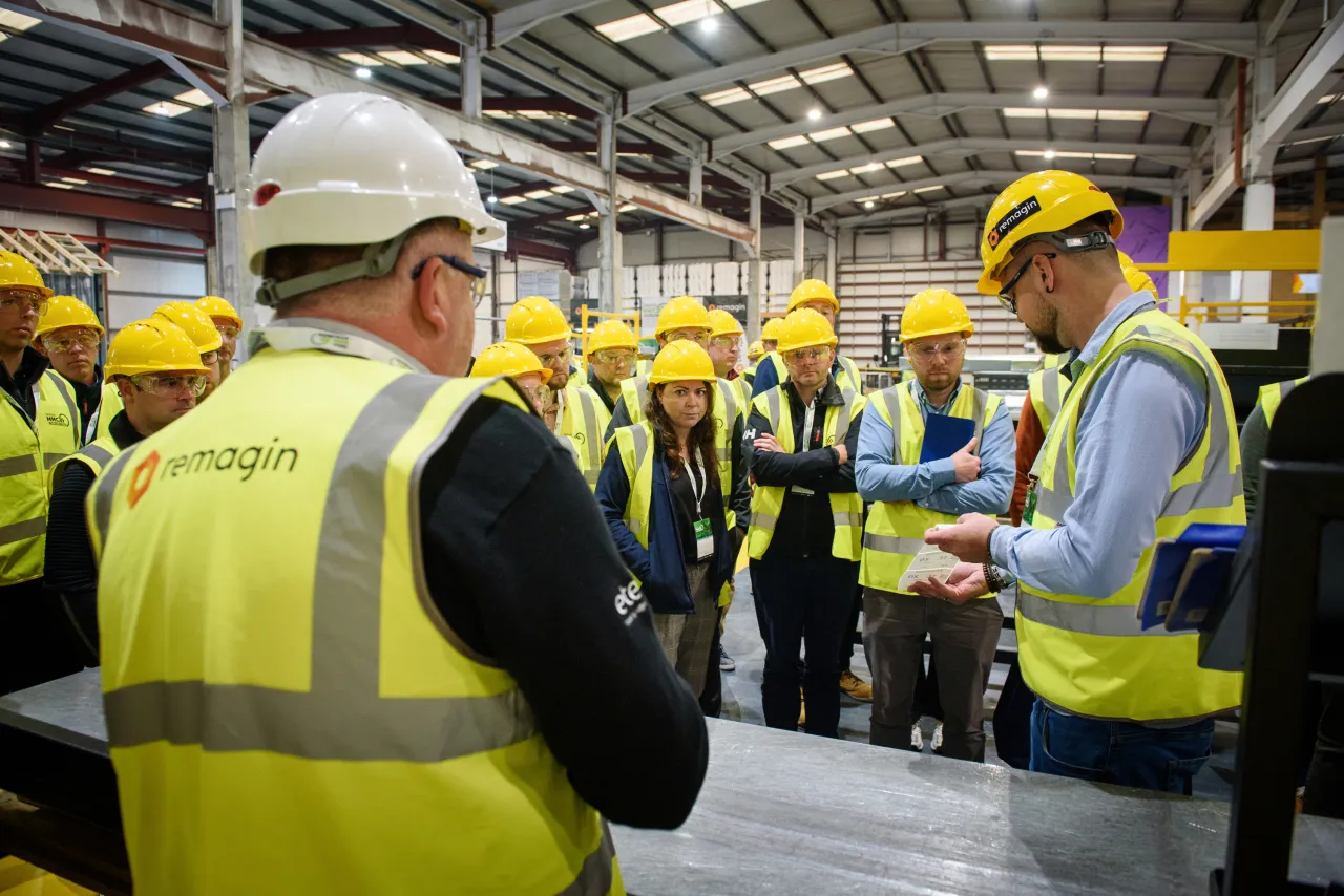 Group of workers wearing yellow hard hats and safety vests gathered in a large industrial facility.