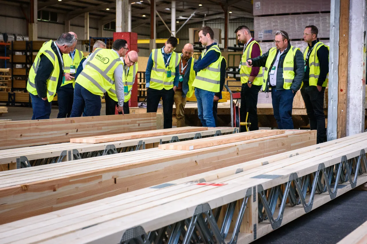 Group of workers in hi-vis jackets inspecting timber beams at a manufacturing facility.