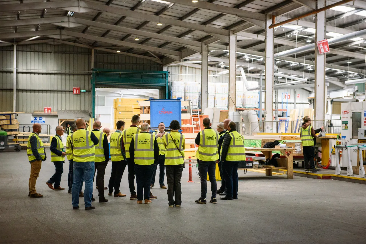 Workers in reflective safety vests gathered in a large industrial warehouse with machinery visible in the background.