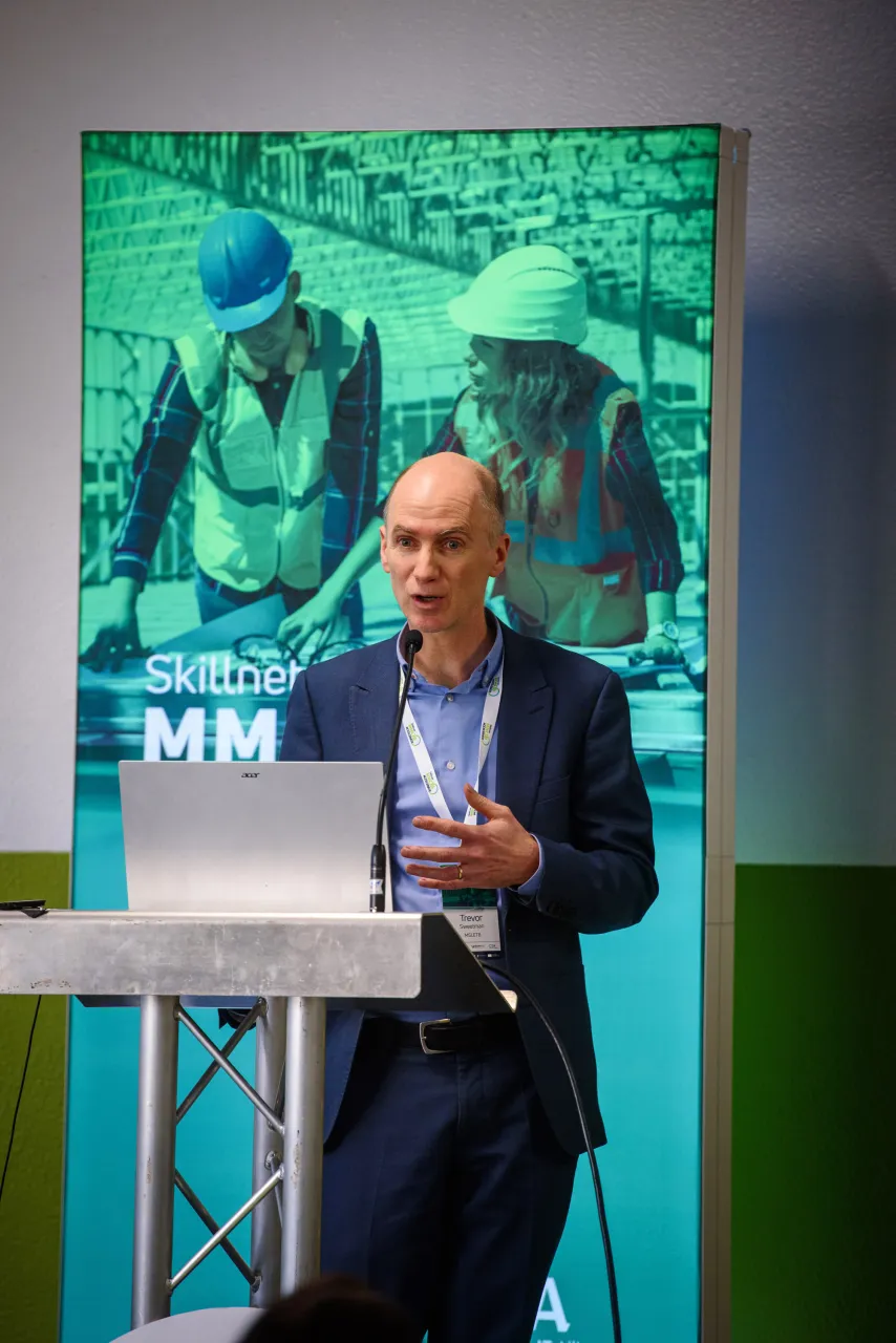 A man in a suit speaking at a presentation with a green-tinted image displayed on screen behind him.