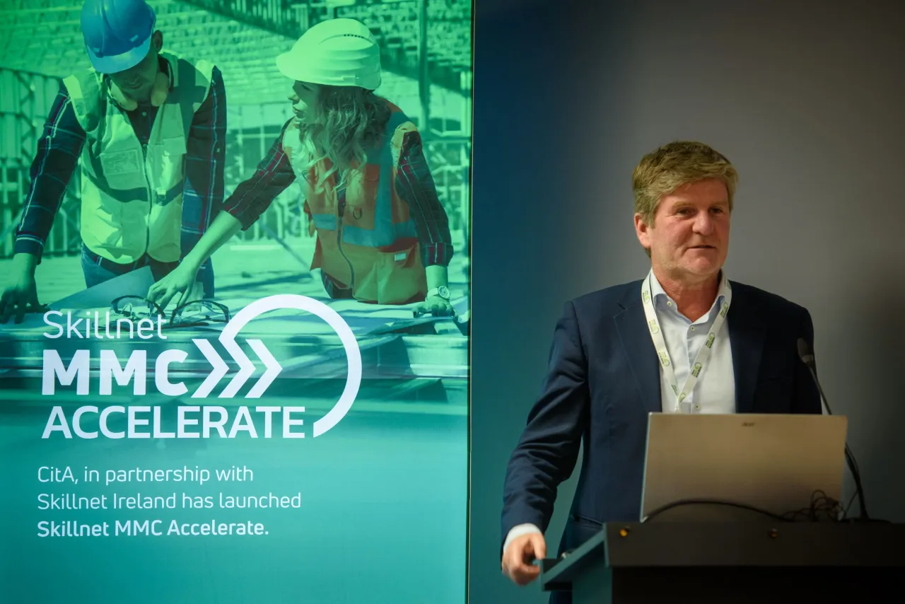 A person standing at podium with green projection screen showing MMEX Accelerate program in background.