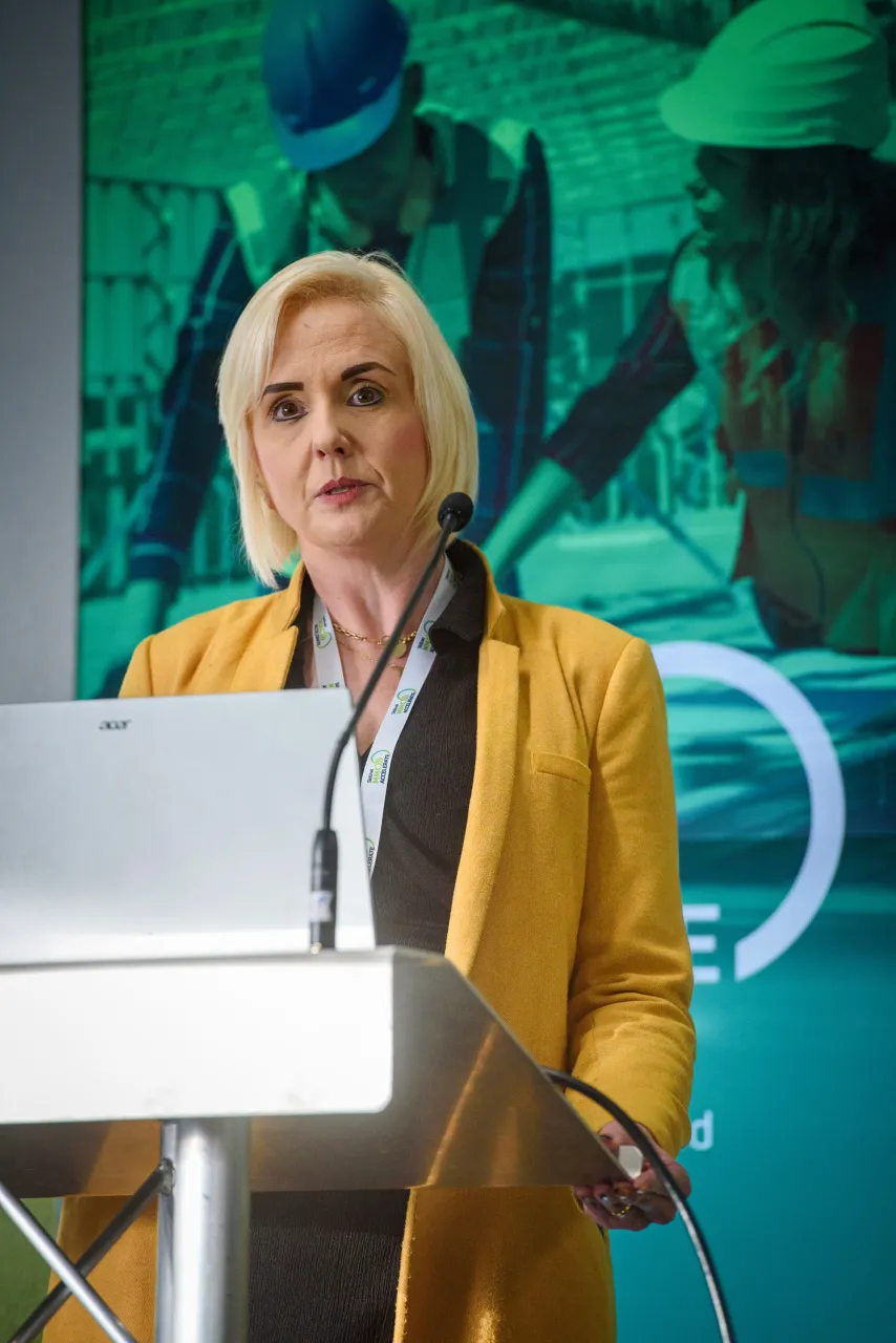A person in a yellow blazer standing at a podium in front of a green backdrop.