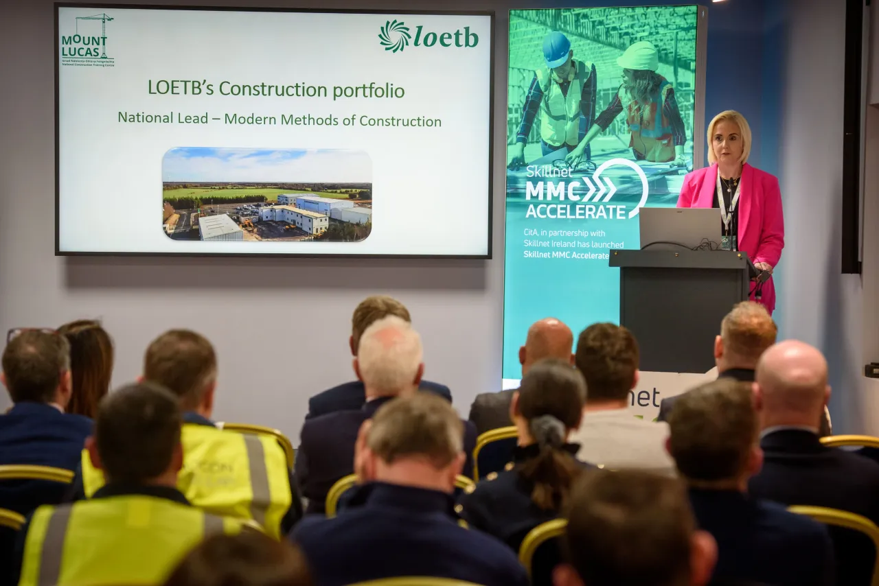 A presenter in a red jacket speaking at a conference with audience members and presentation slides visible.