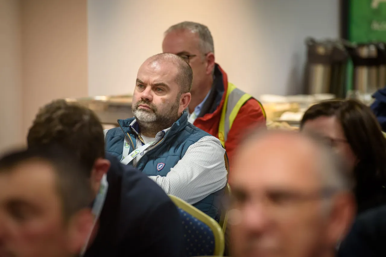 A person with a beard in business attire, sitting among audience members at an event.