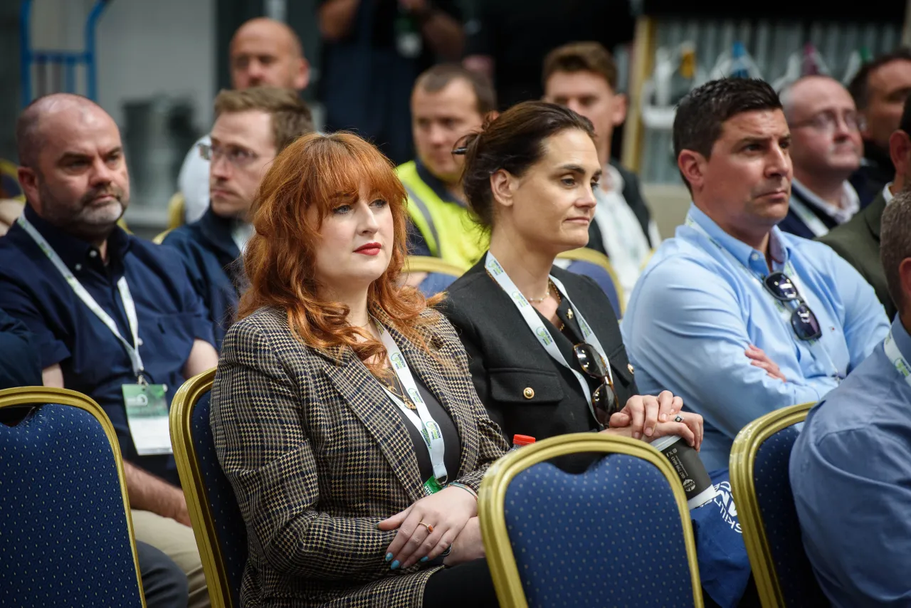 Conference attendees seated in a row, focused on a presentation in a formal setting.