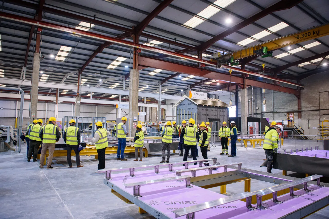 A large warehouse space with workers in safety vests operating manufacturing equipment and handling materials.